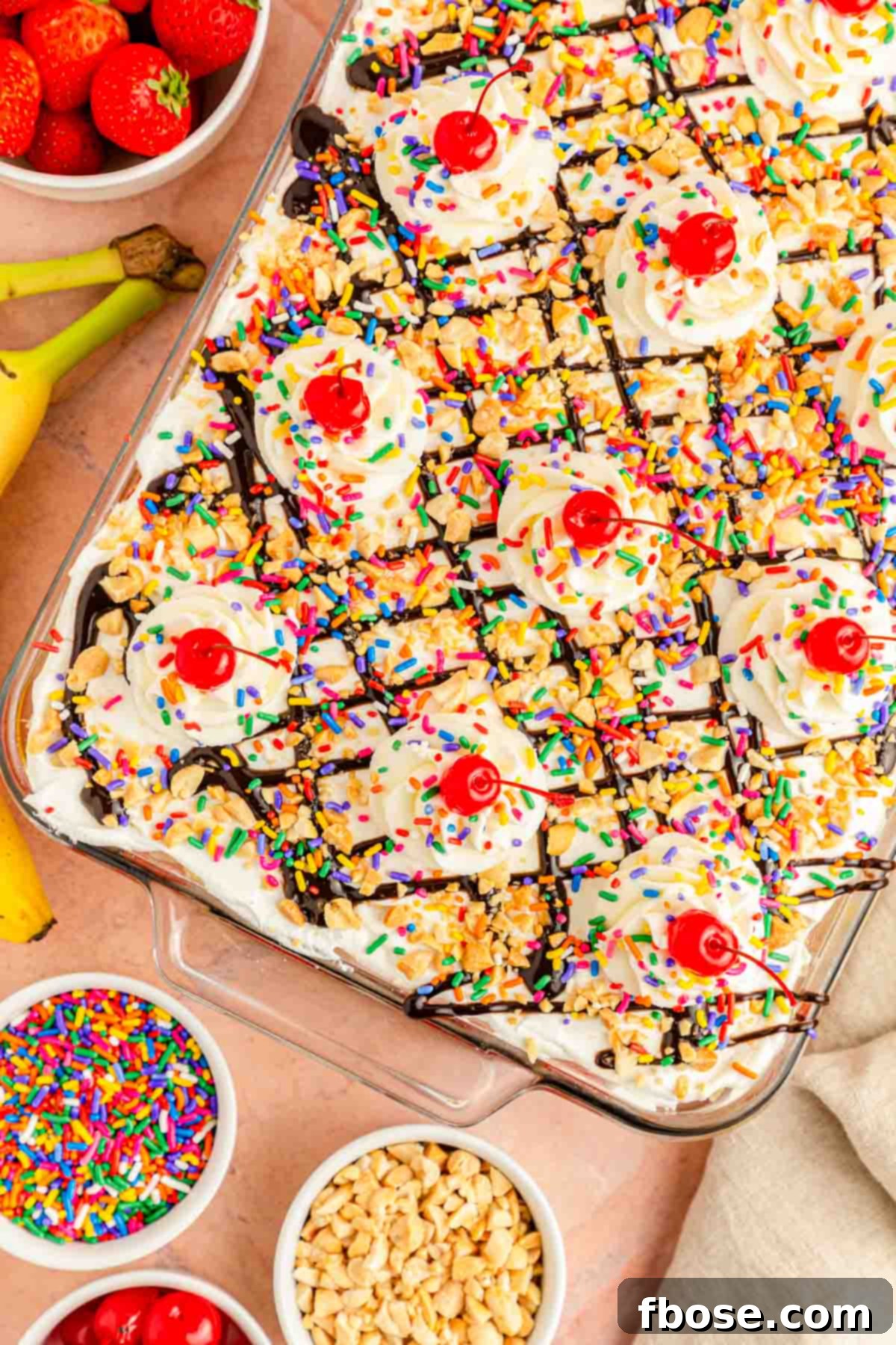 The fully assembled no-bake banana split icebox cake, topped with whipped cream, chocolate drizzle, nuts, sprinkles, and cherries, sitting on a pink table.