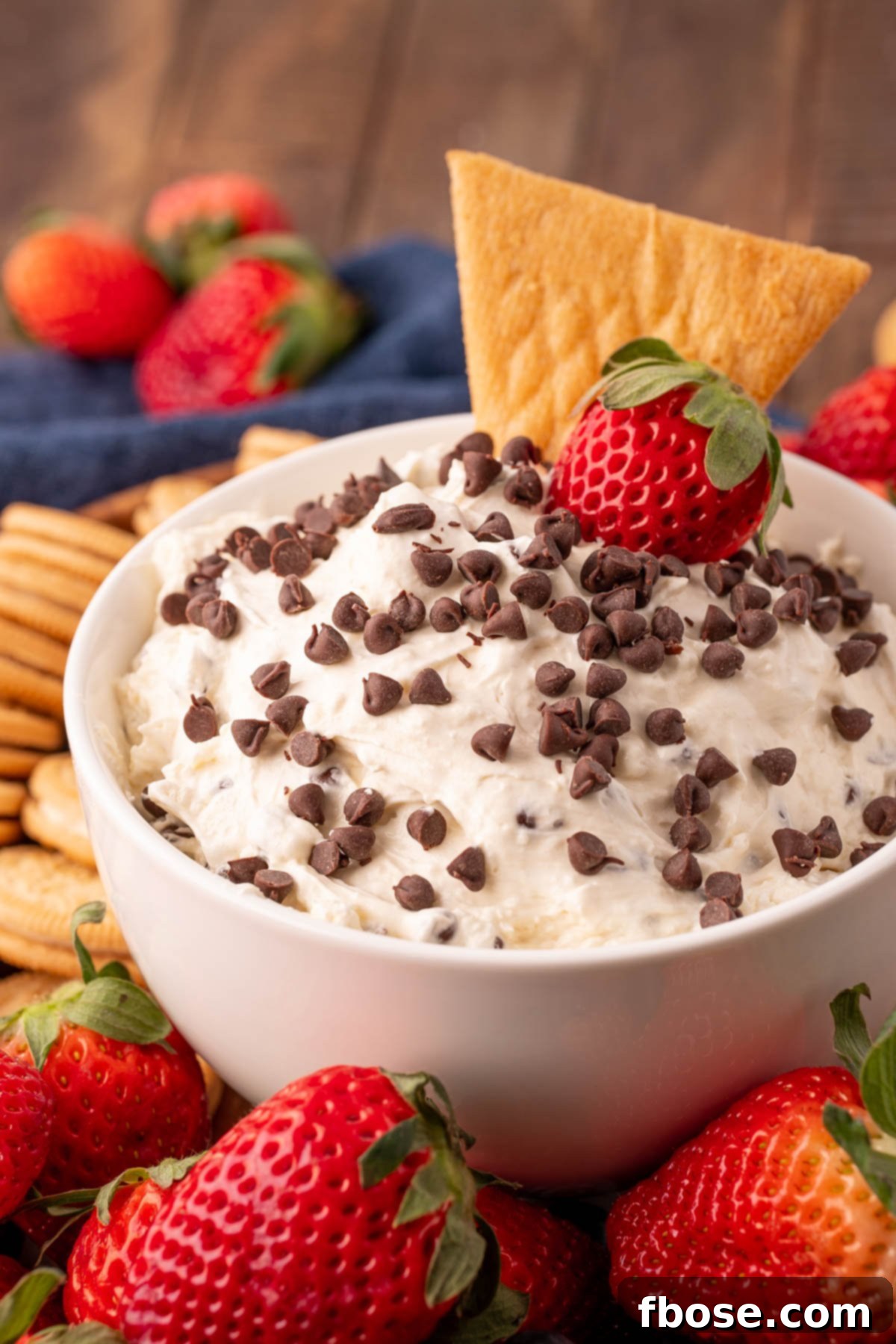Close up of a creamy Booty Dip in a white bowl, surrounded by fresh fruit slices and assorted cookies perfect for dipping. The texture is smooth and inviting, with visible chocolate chips.