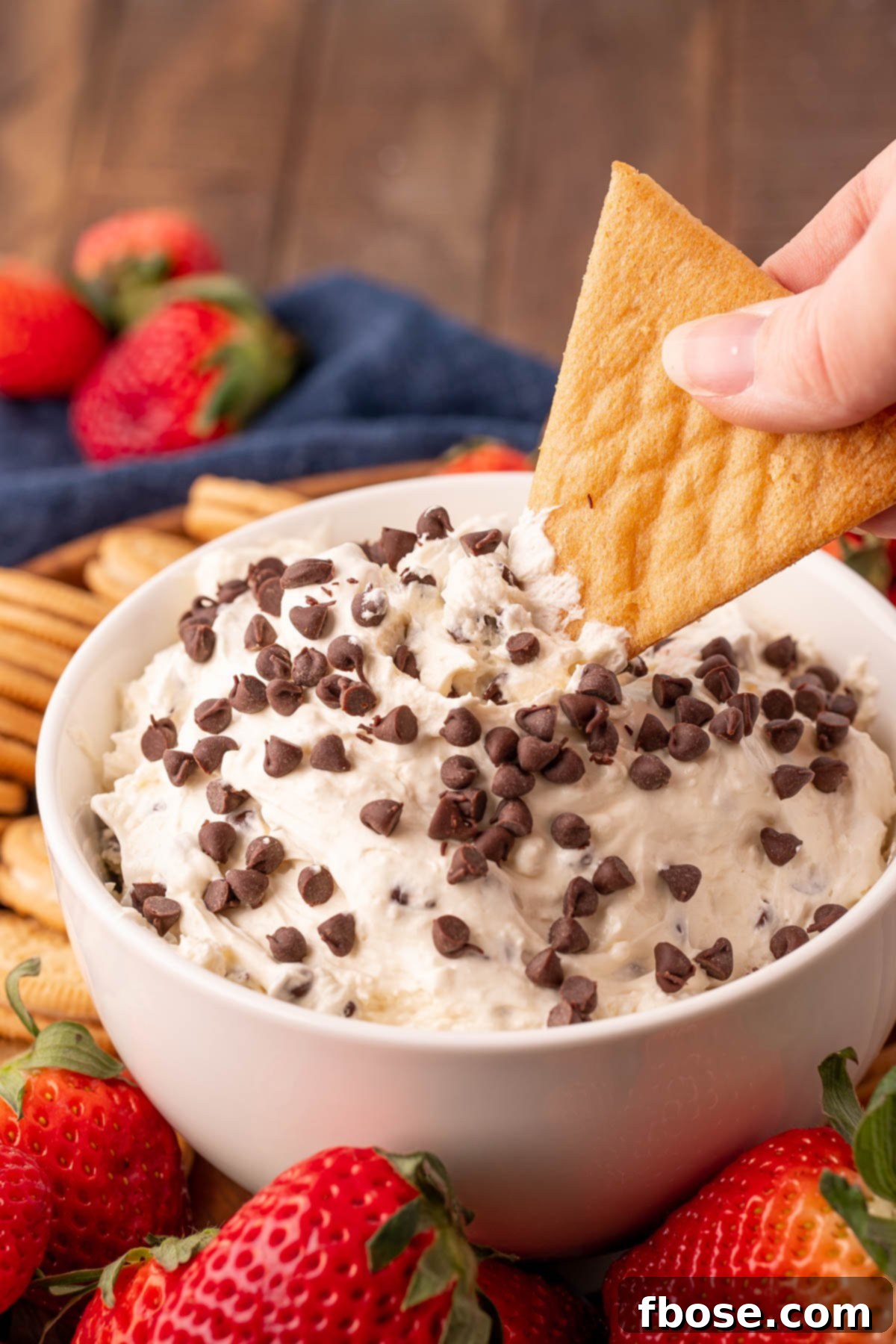 A woman's hand gracefully dips a golden-brown cookie into a creamy white bowl of Booty Dip, showcasing the smooth texture and delicious appeal of the dessert.
