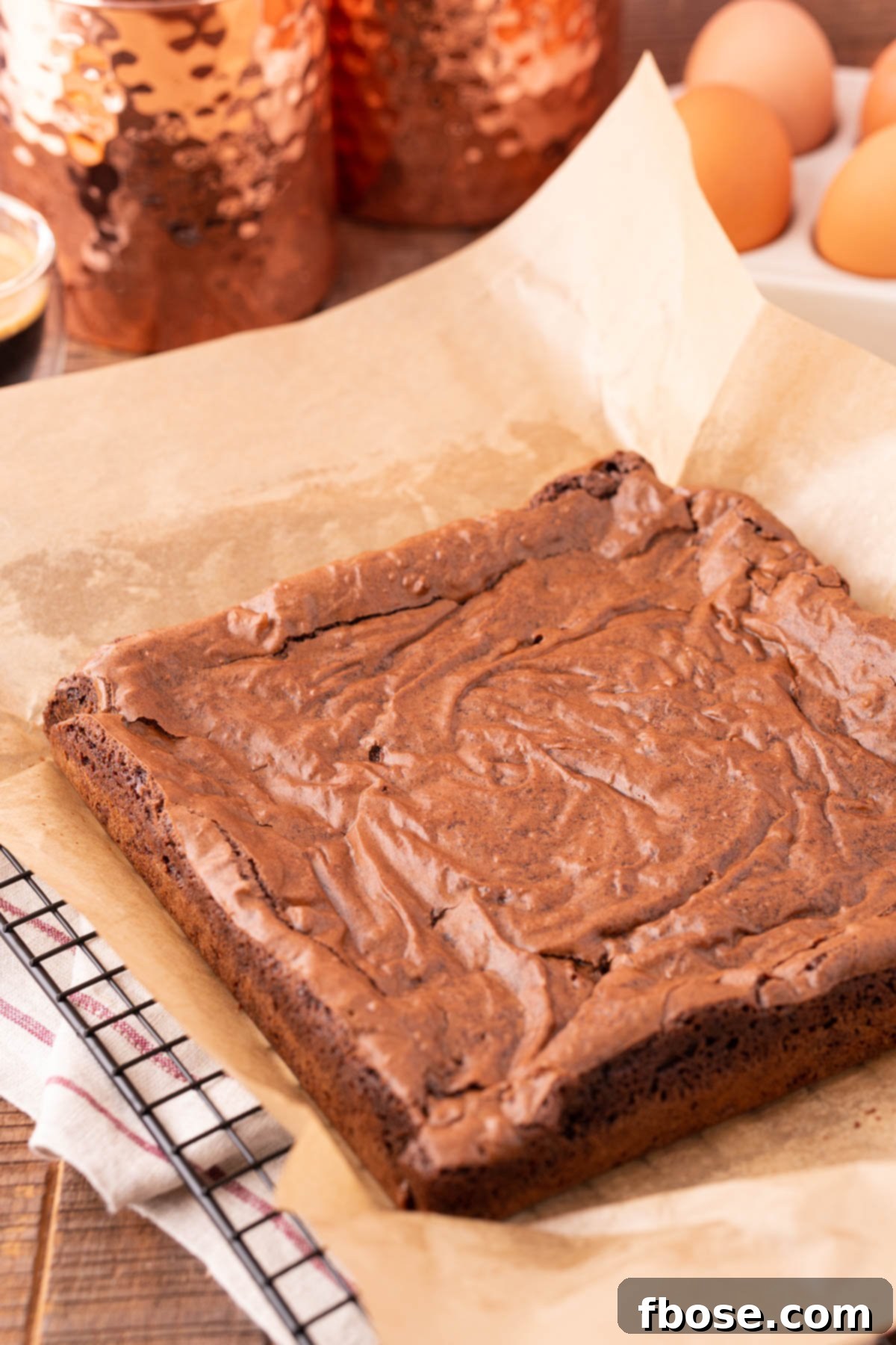 Freshly baked brownies, glistening with a perfect crackly top, resting on a sheet of parchment paper, ready to be cut.