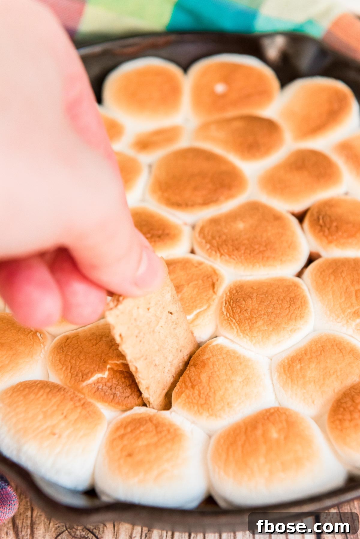 Woman's hand scooping a graham cracker piece into a cast iron skillet filled with toasted marshmallows.