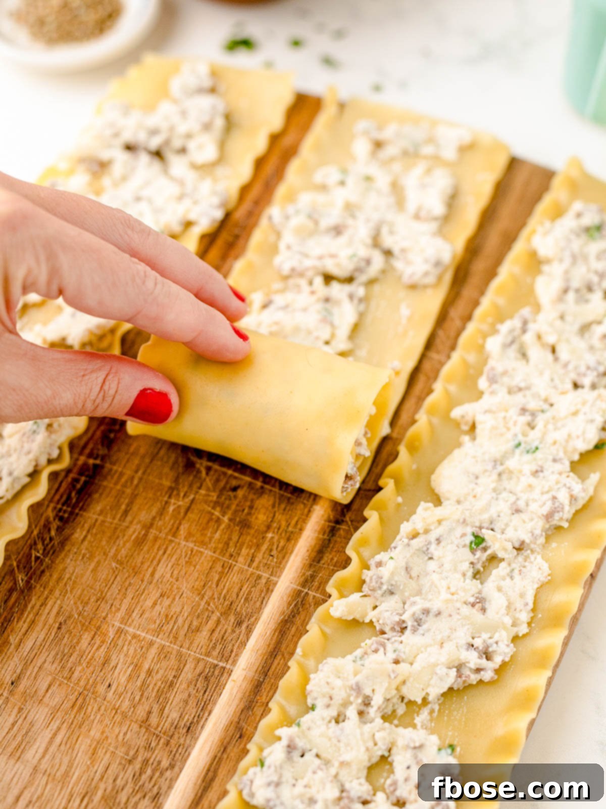 A close-up shot of a woman's hands carefully rolling a lasagna noodle filled with a rich cheese mixture, illustrating the hands-on preparation process.