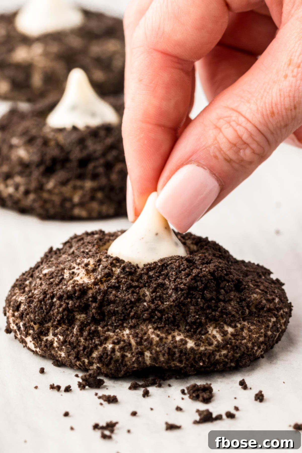 A Cookies and Cream Hershey Kiss being gently pressed into a warm, Oreo crumb-covered cookie, creating the perfect thumbprint.