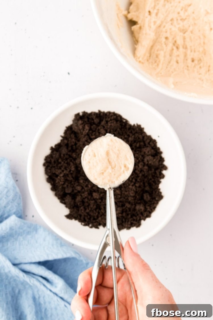 A cookie scoop with sugar cookie dough poised over a bowl of crushed Oreos, ready for coating.