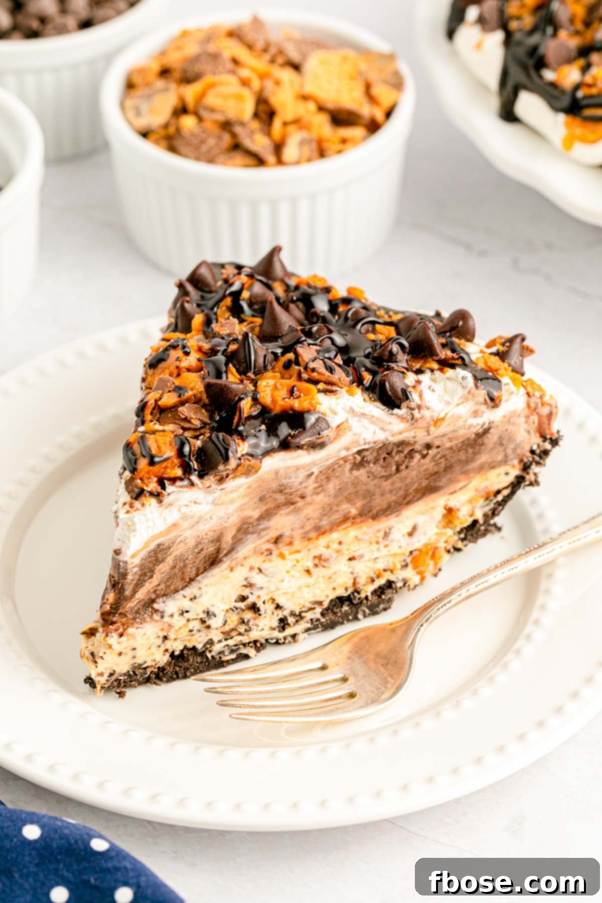 A close up of a perfectly sliced No Bake Butterfinger Pie, showcasing its distinct layers of creamy peanut butter, rich chocolate fudge, and a crunchy Oreo crust, generously topped with Butterfinger candy pieces, resting on a pristine white plate.