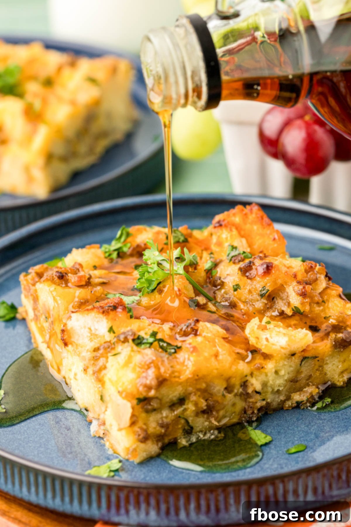 Pure maple syrup being drizzled generously over a warm slice of Overnight Breakfast Sausage Strata on a plate.