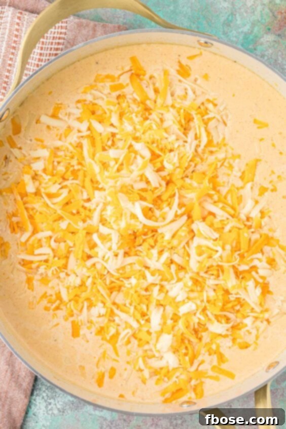 Shredded cheese being gradually added to a creamy sauce in a skillet, melting into the mac and cheese base.