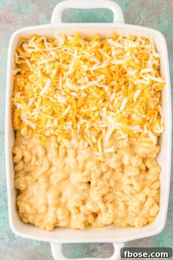 Mac and cheese being layered with additional shredded cheese in a baking dish before baking.