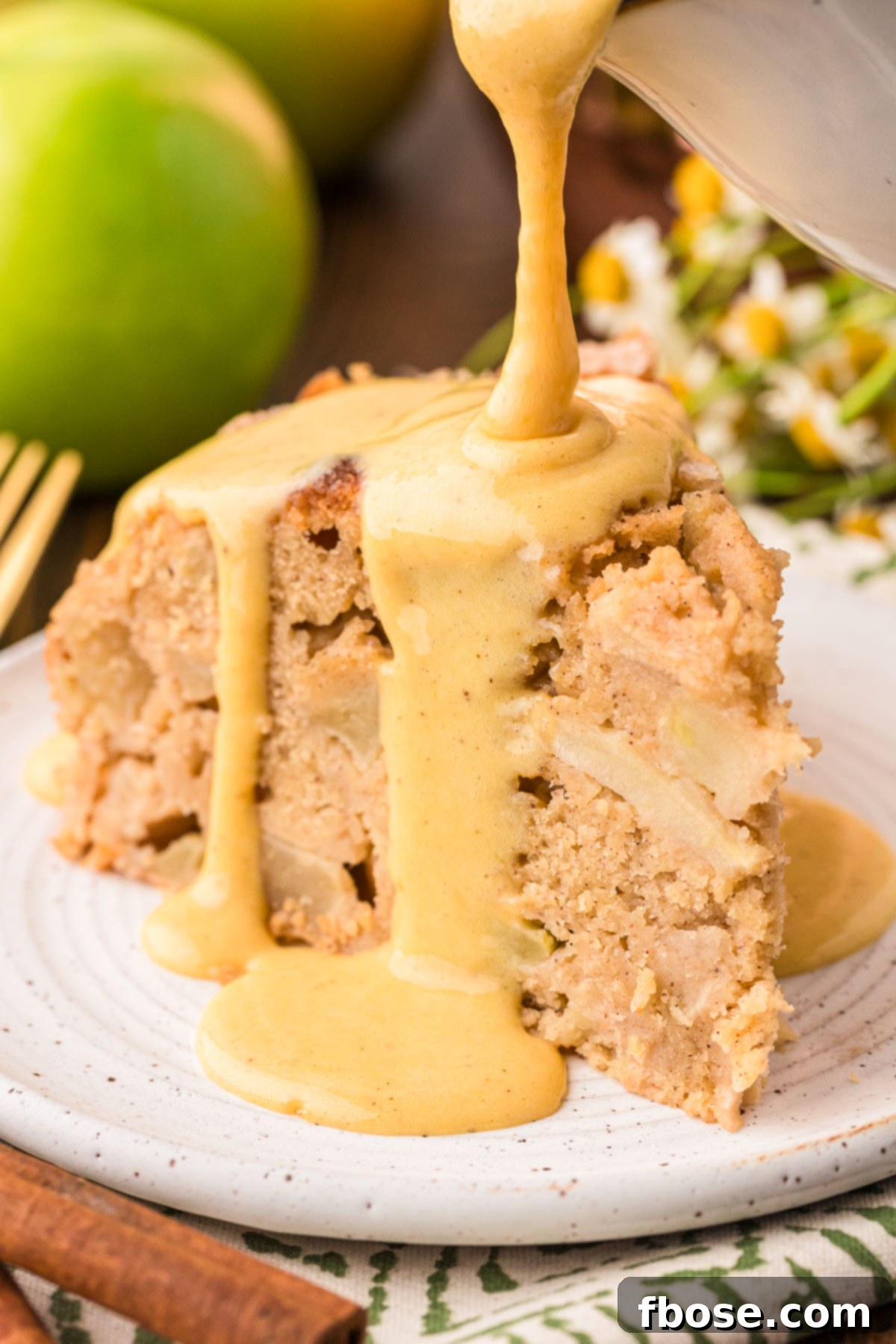Vanilla custard being gracefully poured over a slice of Irish Apple Cake on a white plate, creating an enticing, glossy finish.