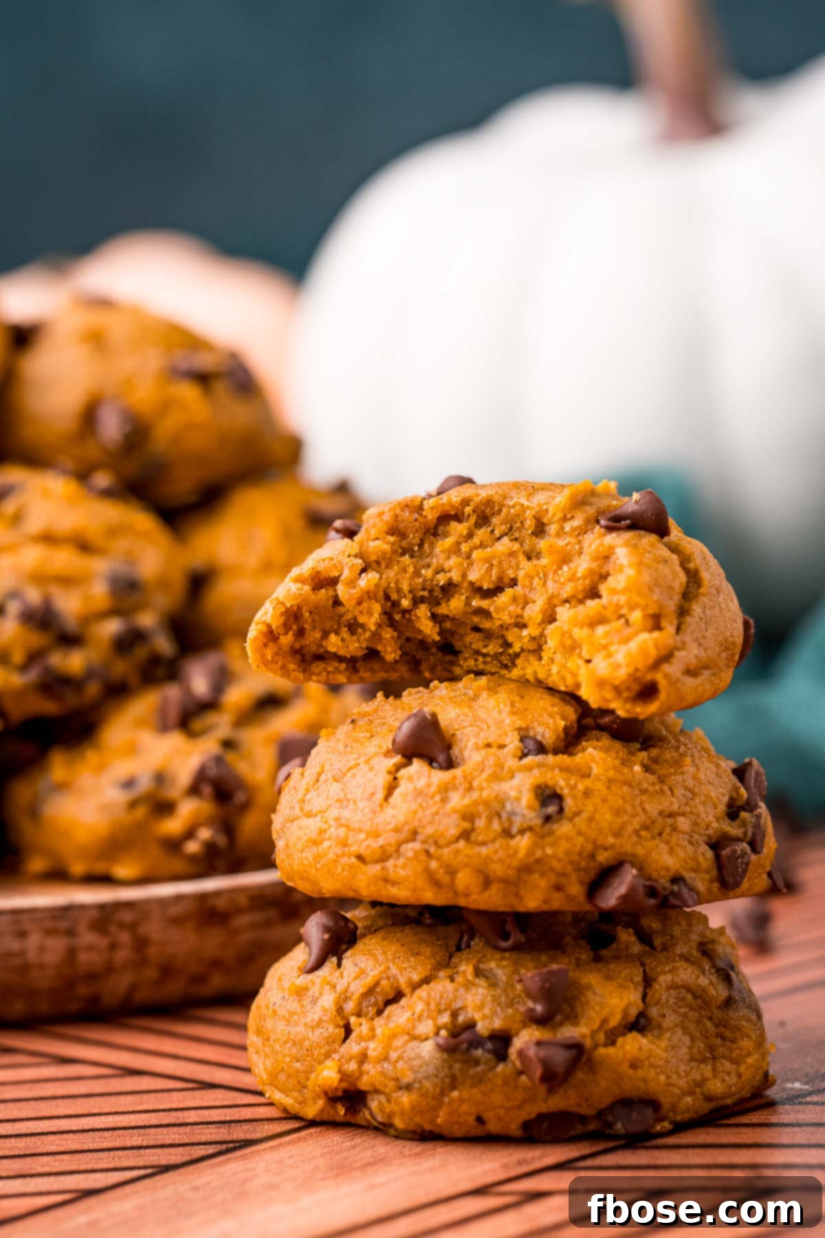 A stack of pumpkin chocolate chip cookies on a table with a bite taken out of the top on. A plate with more cookies is in the background.