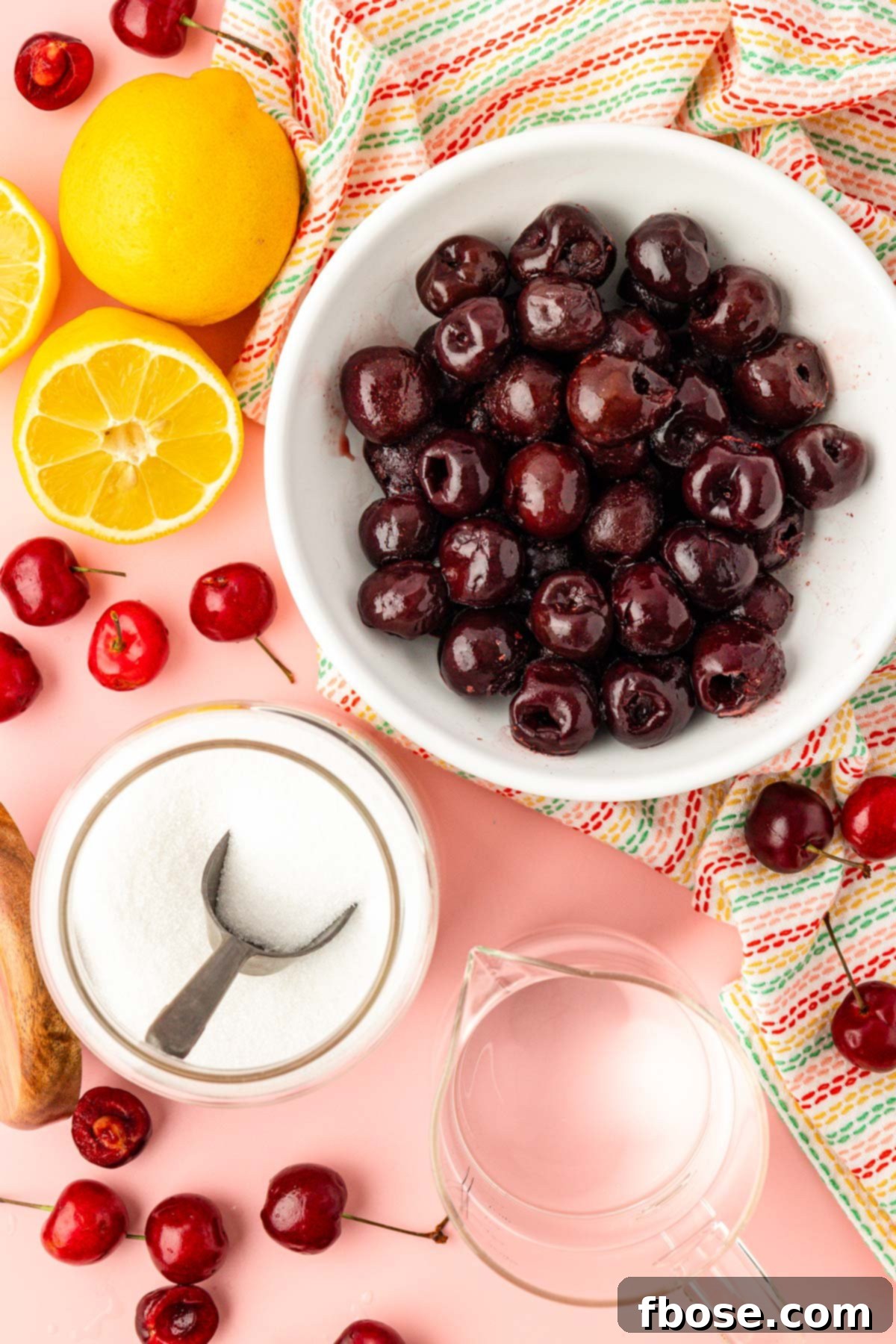 An inviting overhead shot showcasing the simple, wholesome ingredients required for cherry simple syrup: vibrant red cherries, bright lemons, clear water, and sparkling granulated sugar, laid out on a clean surface.