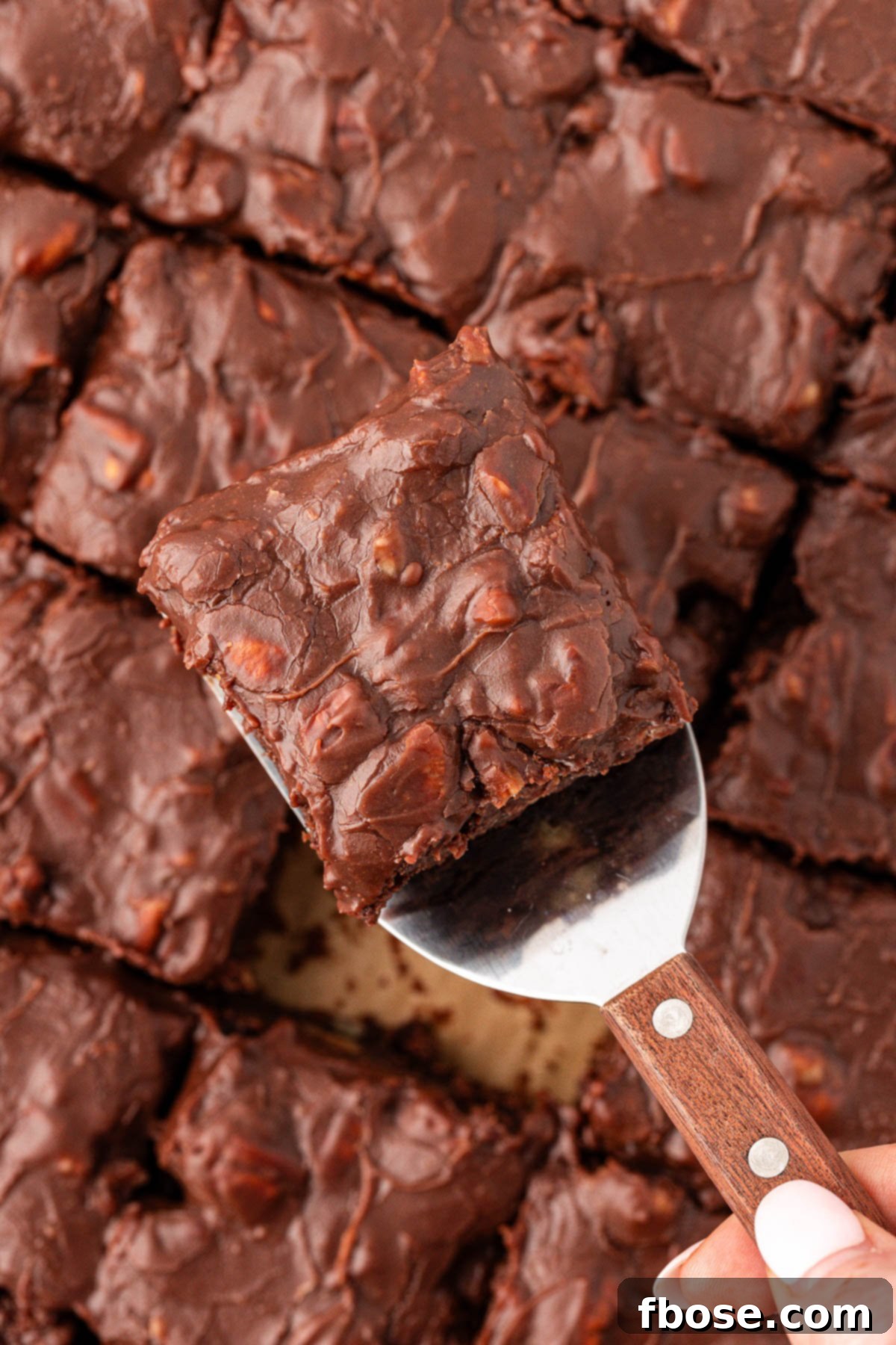 Overhead photo of a brownie topped with icing and pecans being lifted out of a pan with the parchment paper, demonstrating easy removal.