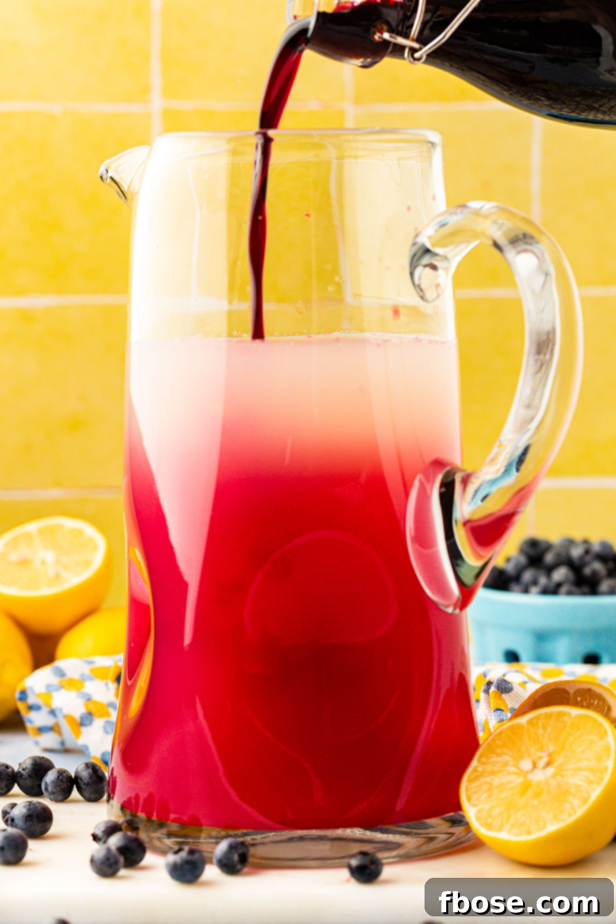 Homemade blueberry lemonade being carefully poured into a large glass pitcher, showcasing its rich purple hue.