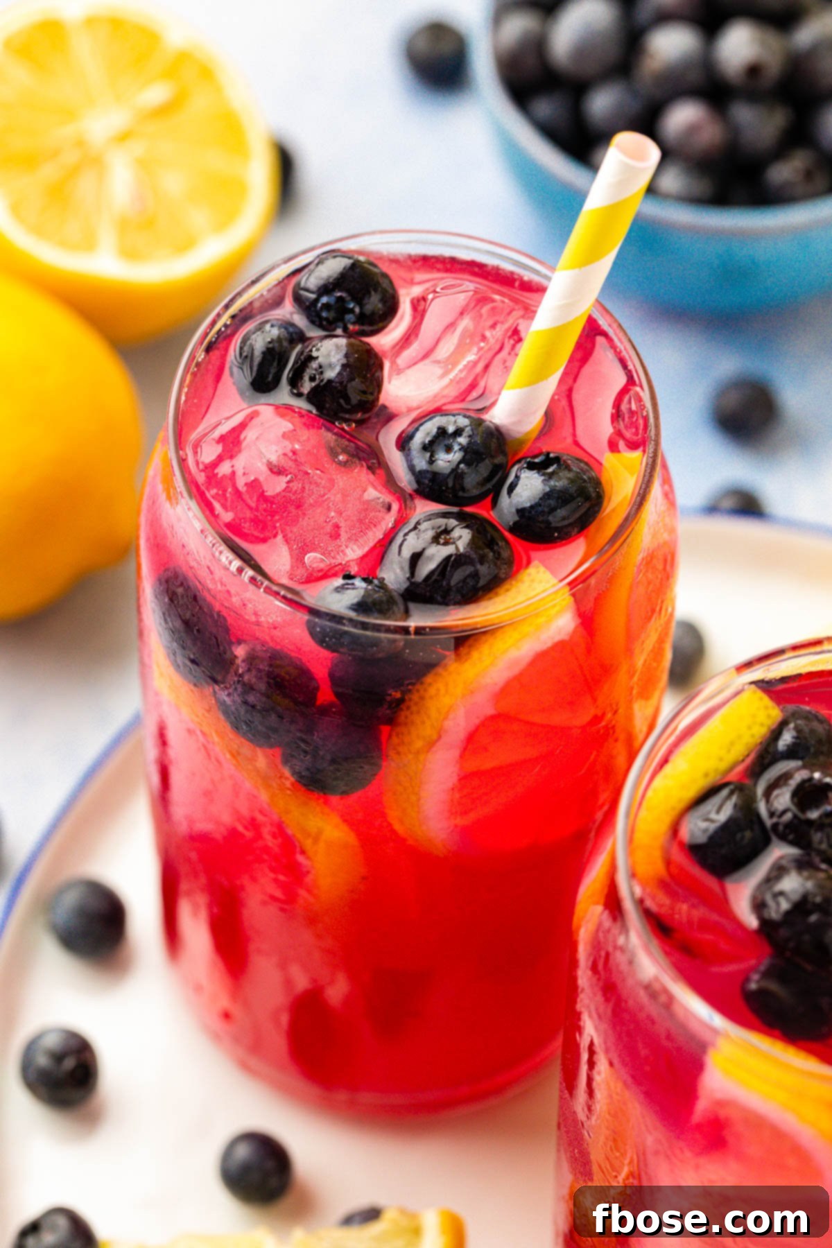 A single glass of blueberry lemonade, garnished with a fresh lemon slice and blueberries, set against a soft, blurred background.