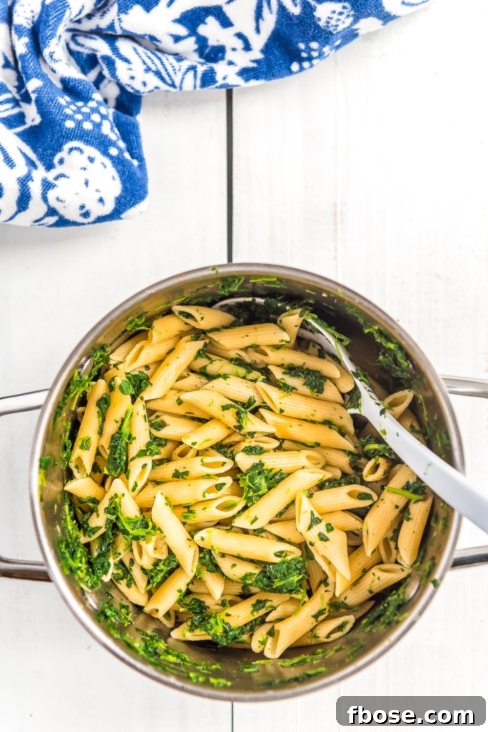 Cooked penne pasta in a pot, mixed with vibrant green spinach, on a white wood table with a blue and white napkin.