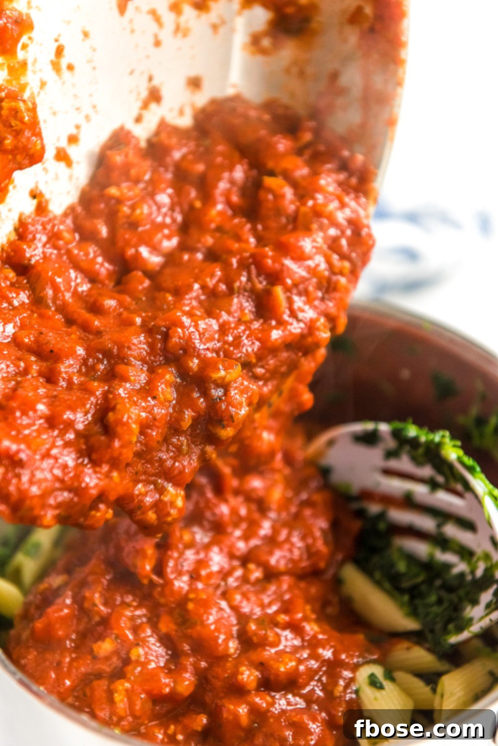 Rich meat sauce being poured into a pot containing cooked pasta and spinach, ready to be mixed.