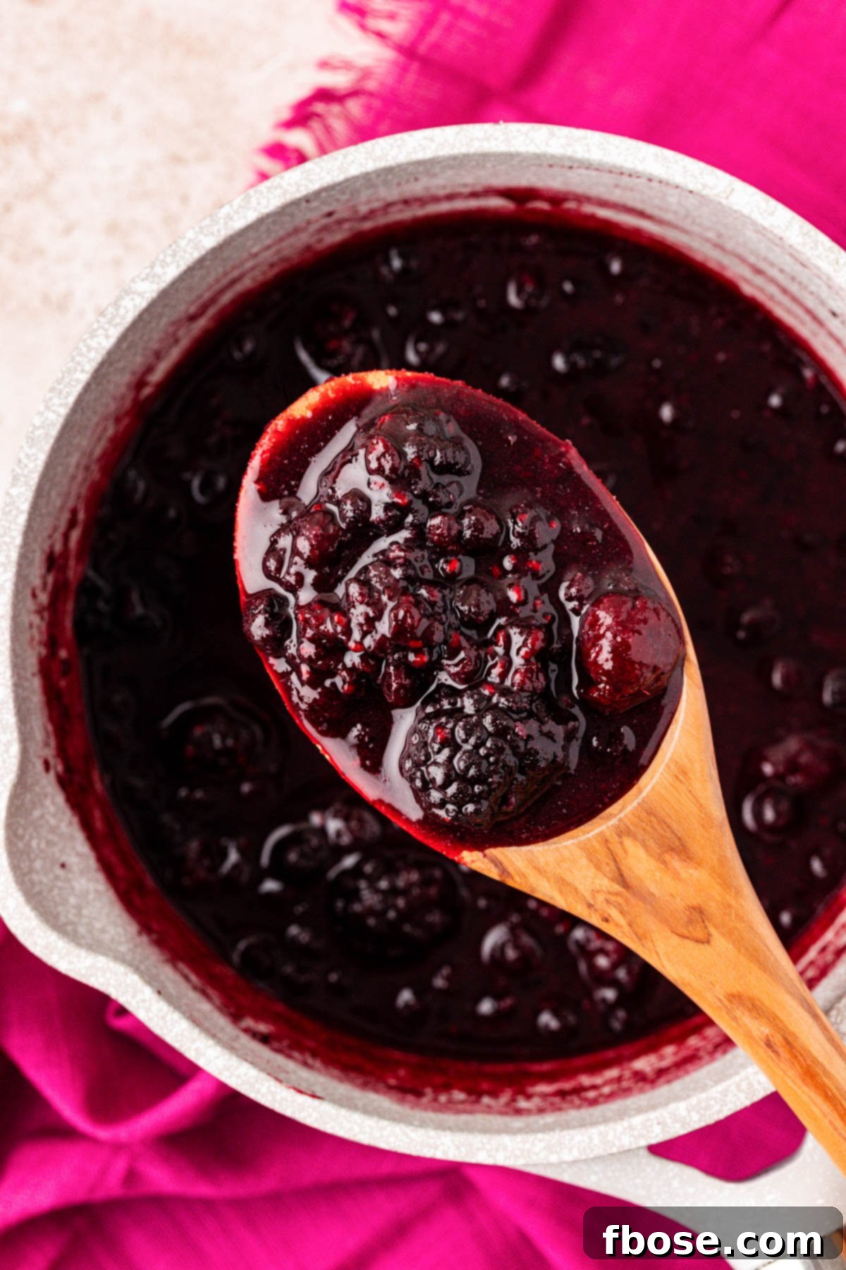 A serving spoon overflows with warm, chunky mixed berry compote, held above the pot from which it was just prepared.