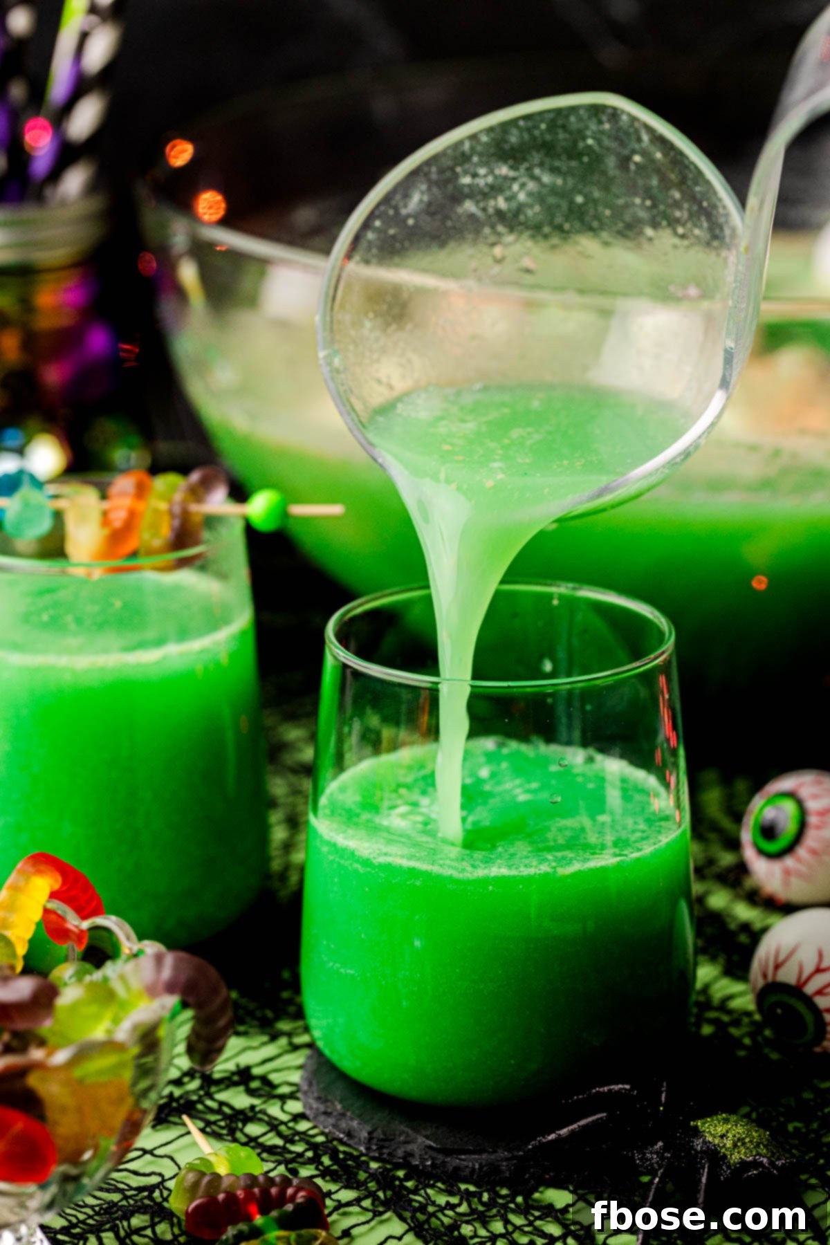 Green Halloween punch being ladled into glasses, showing the frothy sherbet and gummy worms, ready to be served at a party.