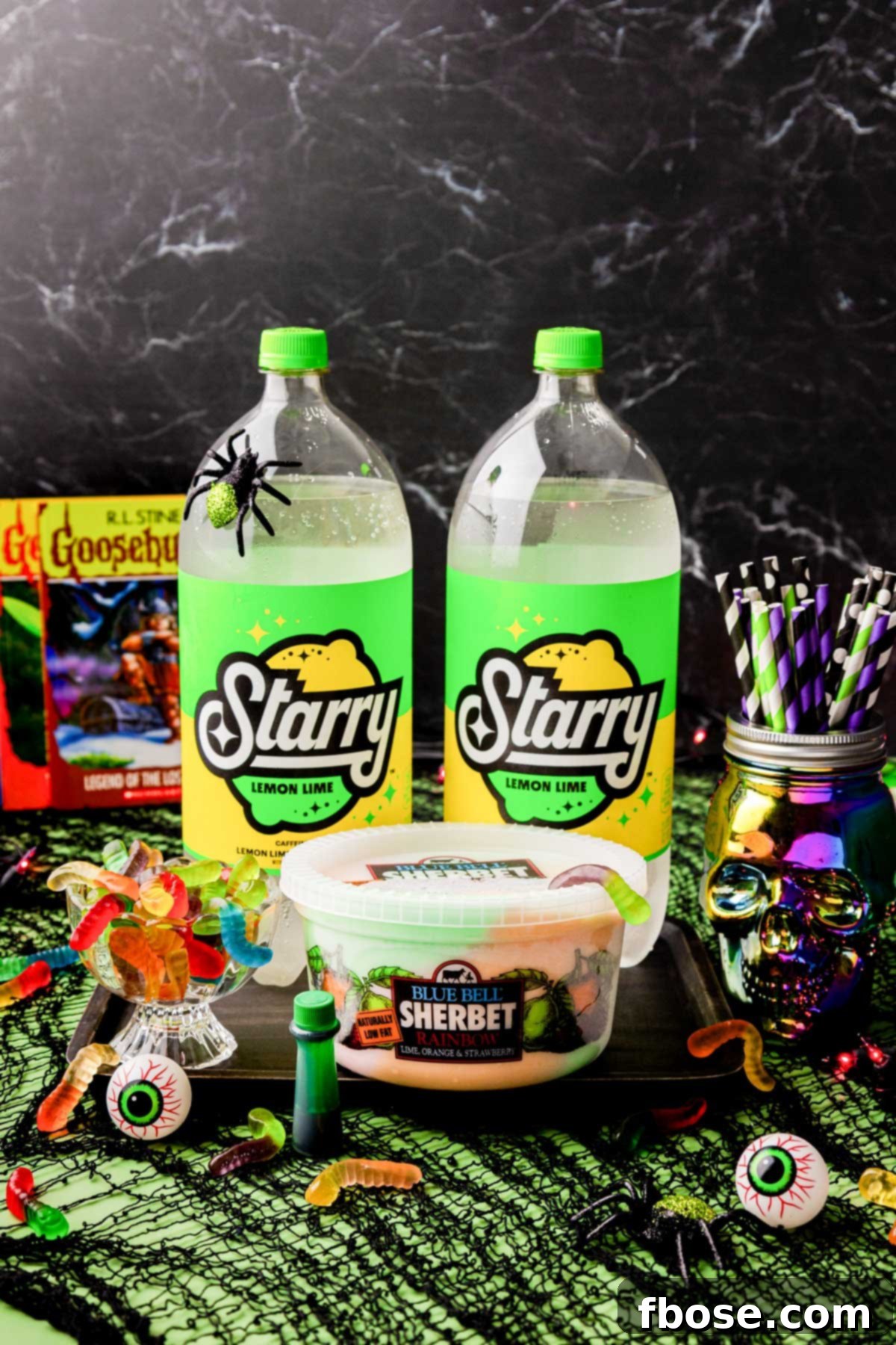 Ingredients laid out on a table for making Halloween punch, including soda, food coloring, sherbet, and gummy worms, all set against a festive party backdrop.
