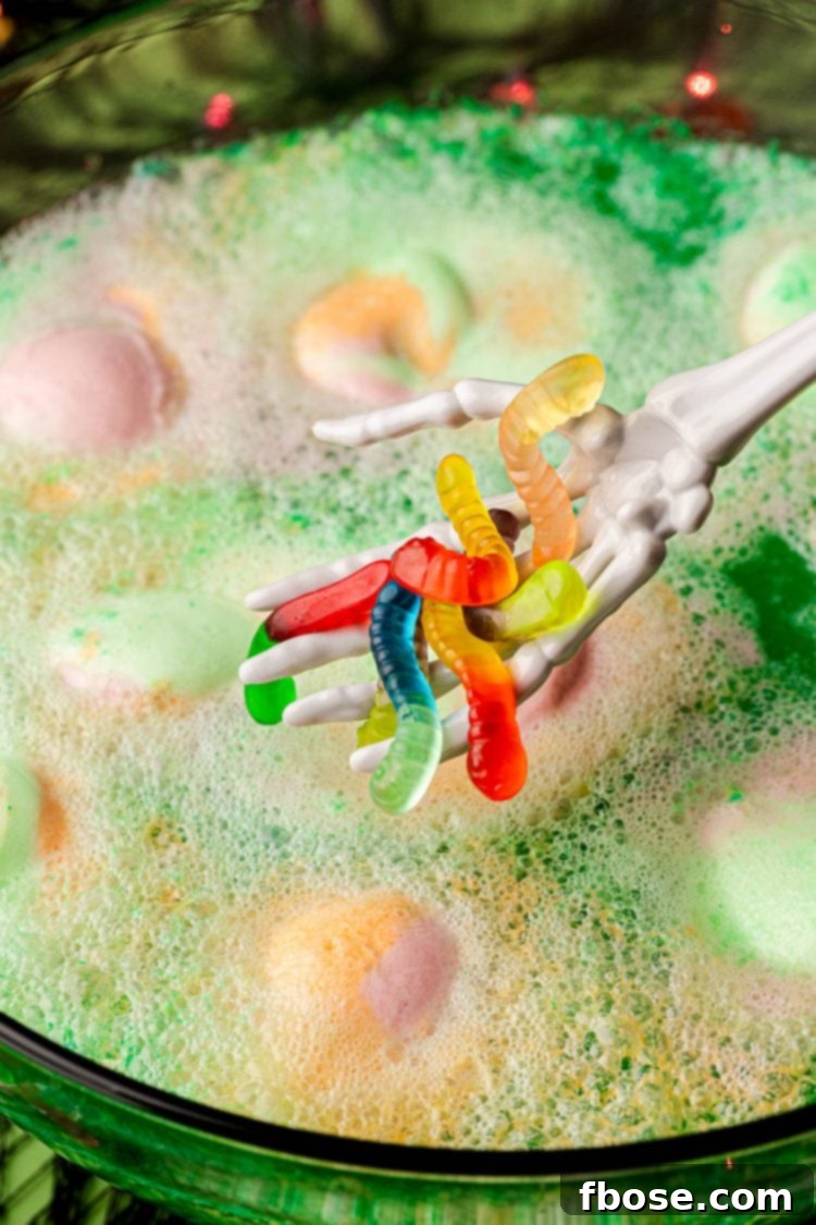 A plastic skeleton hand adding gummy worms to a bowl of green Halloween punch for a spooky effect.
