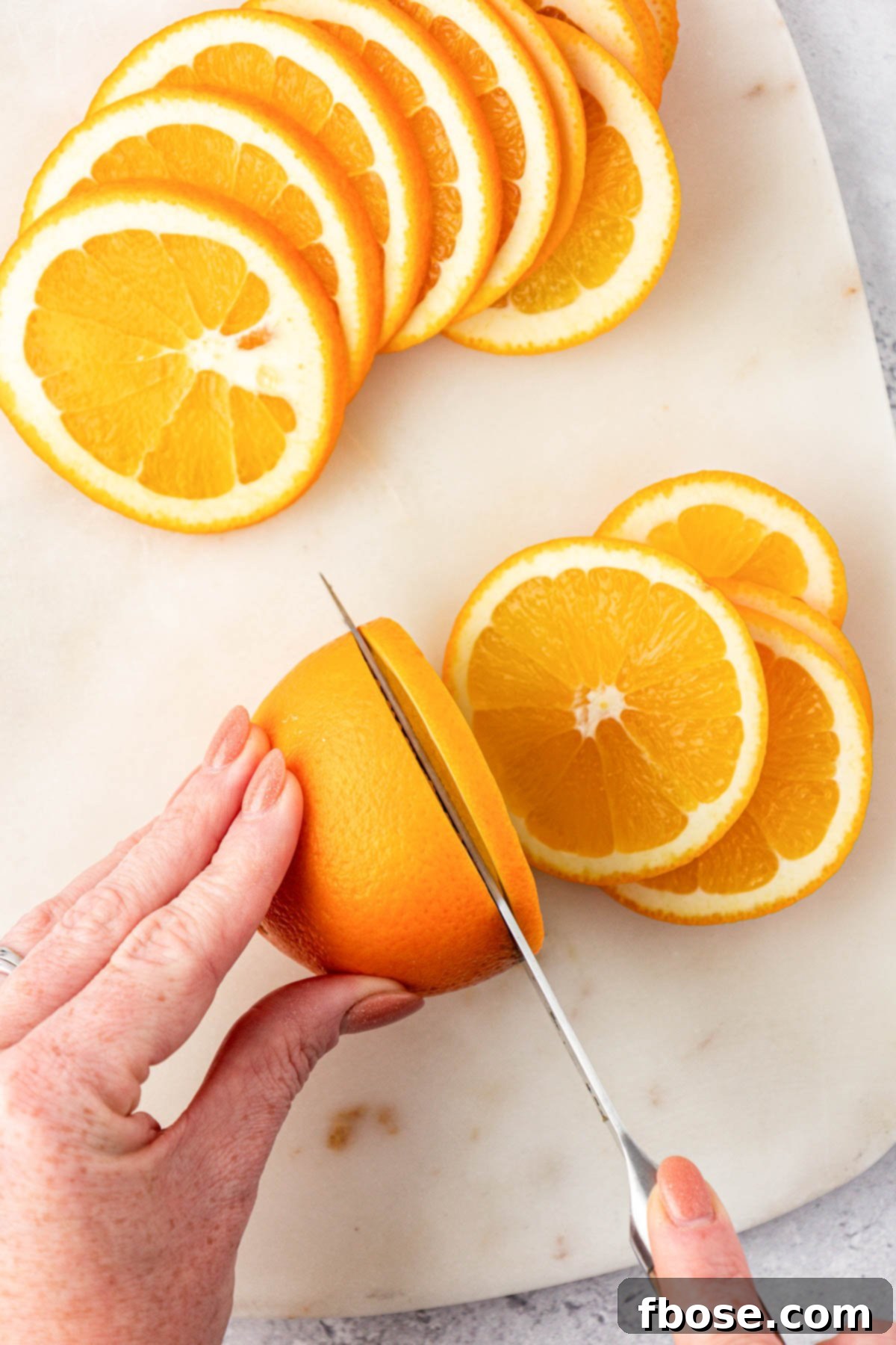 Oven-Dried Orange Slices 4 A woman's hand meticulously cutting navel oranges into thin, uniform slices on a wooden cutting board.