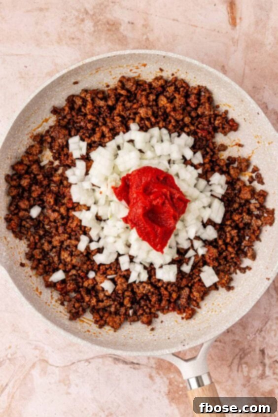 Onion and tomato paste being added to a skillet with ground beef.