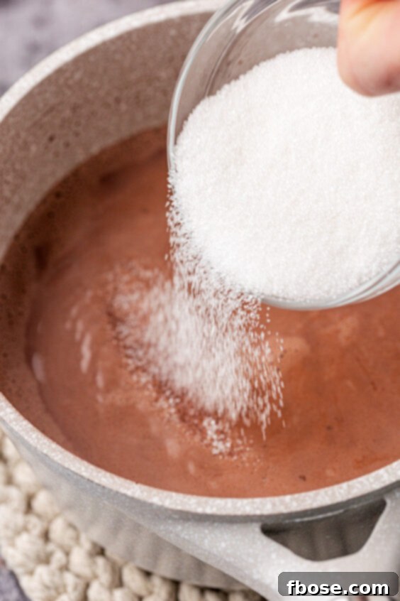 Granulated sugar being poured from a small bowl into the hot chocolate mixture in a pot.
