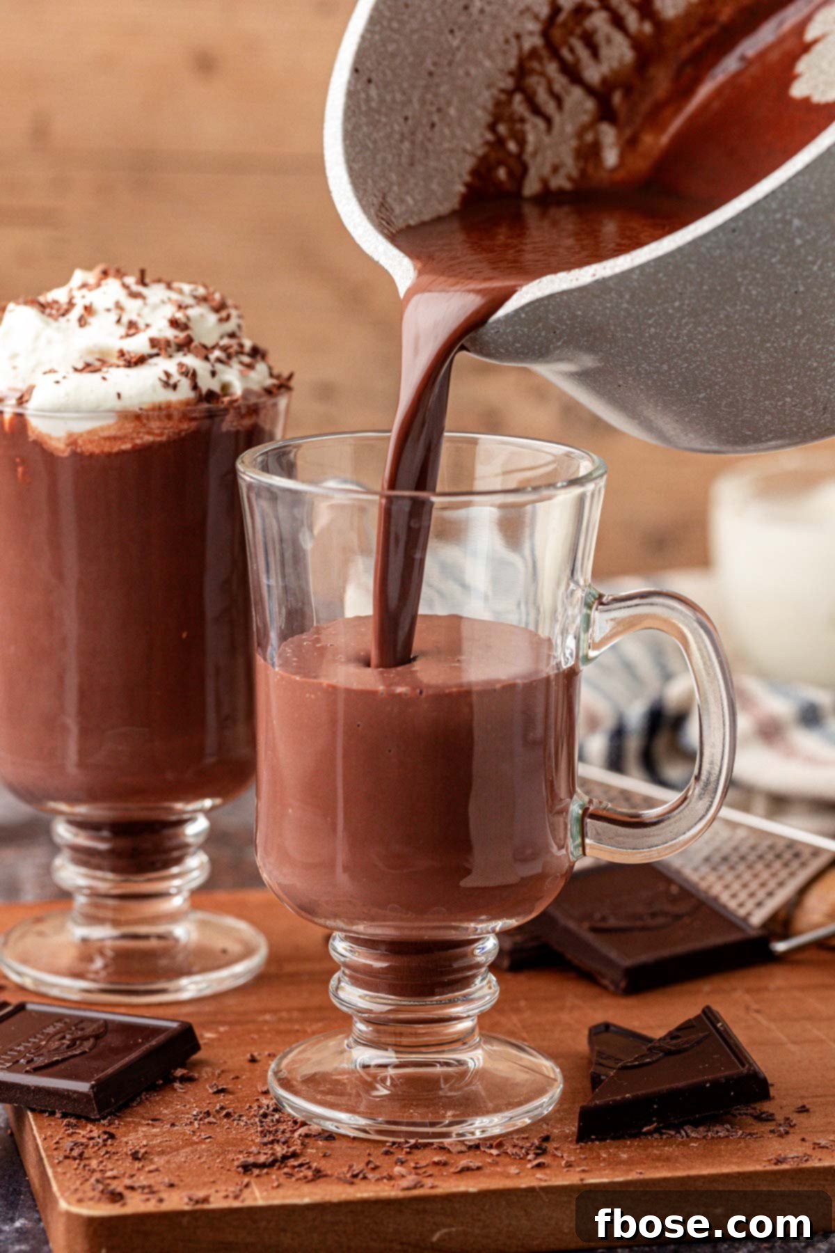 Thick, dark hot chocolate being poured from a saucepan into an elegant glass mug, showcasing its rich texture.