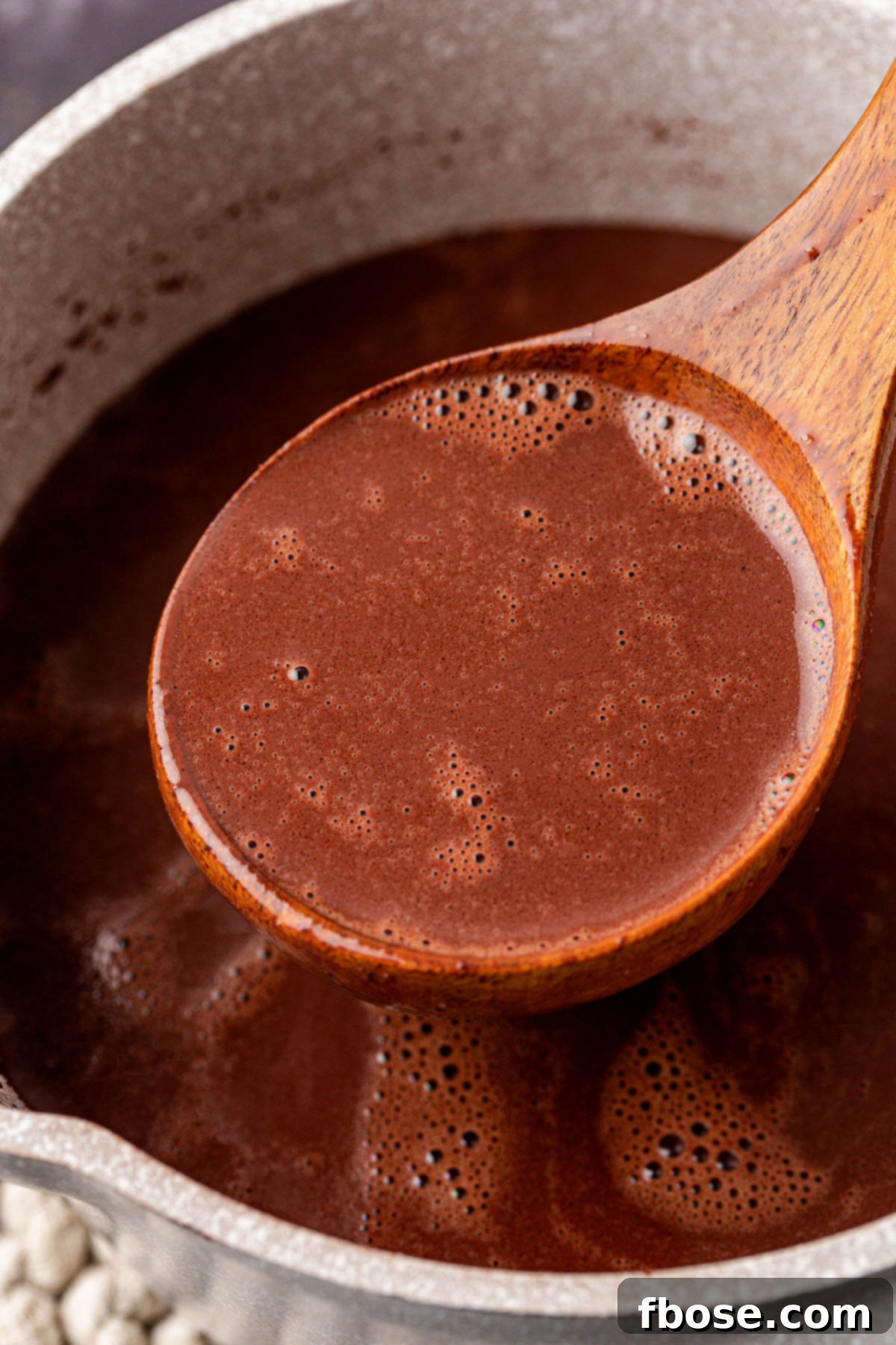 A wooden ladle lifting thick, dark hot chocolate from a copper pot, highlighting its rich, glossy texture.