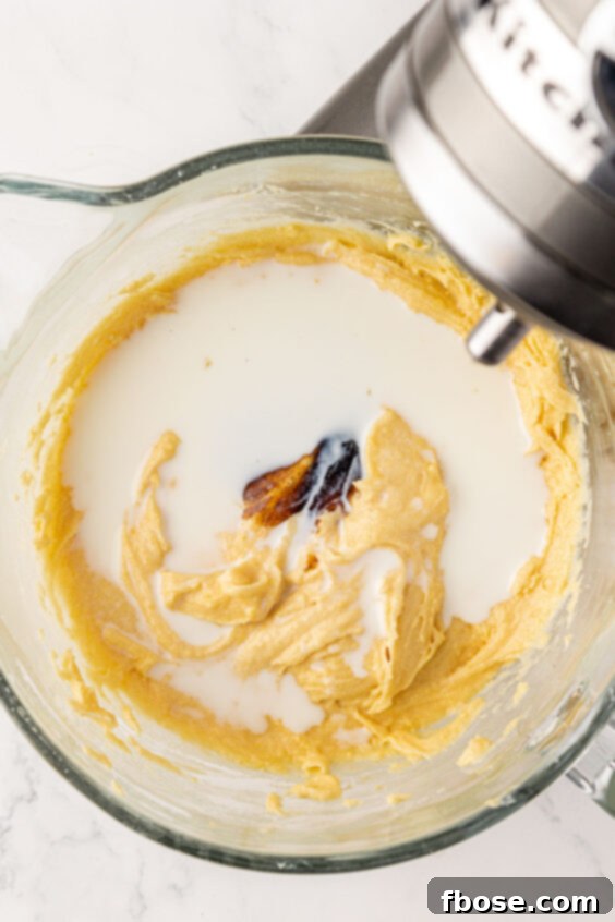 Vanilla bean paste and milk being added to cake batter in a mixing bowl.
