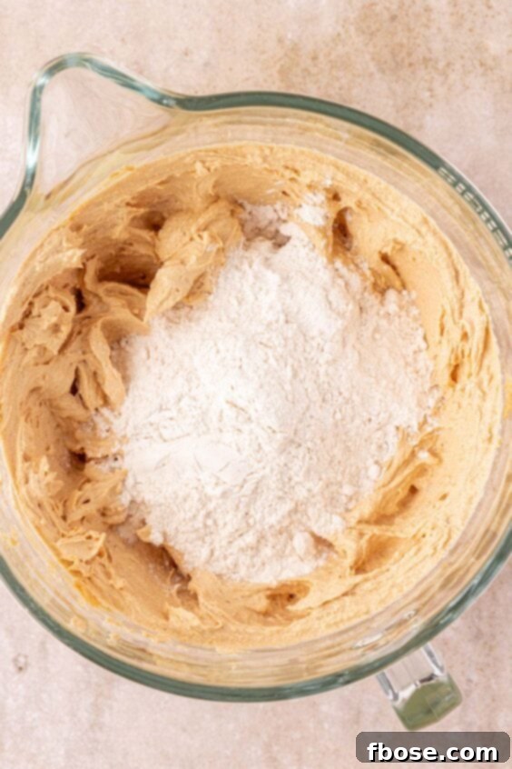 Dry ingredients being added to wet ingredients in a mixing bowl to make cookie dough, showing the gradual mixing process.