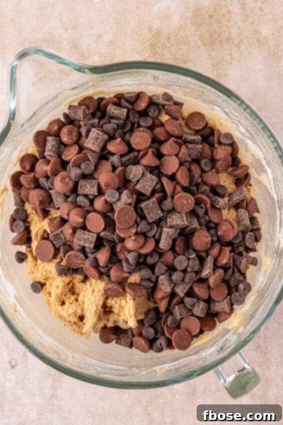 Chocolate chips and chunks being added to cookie dough in a mixing bowl, illustrating the final step before portioning.