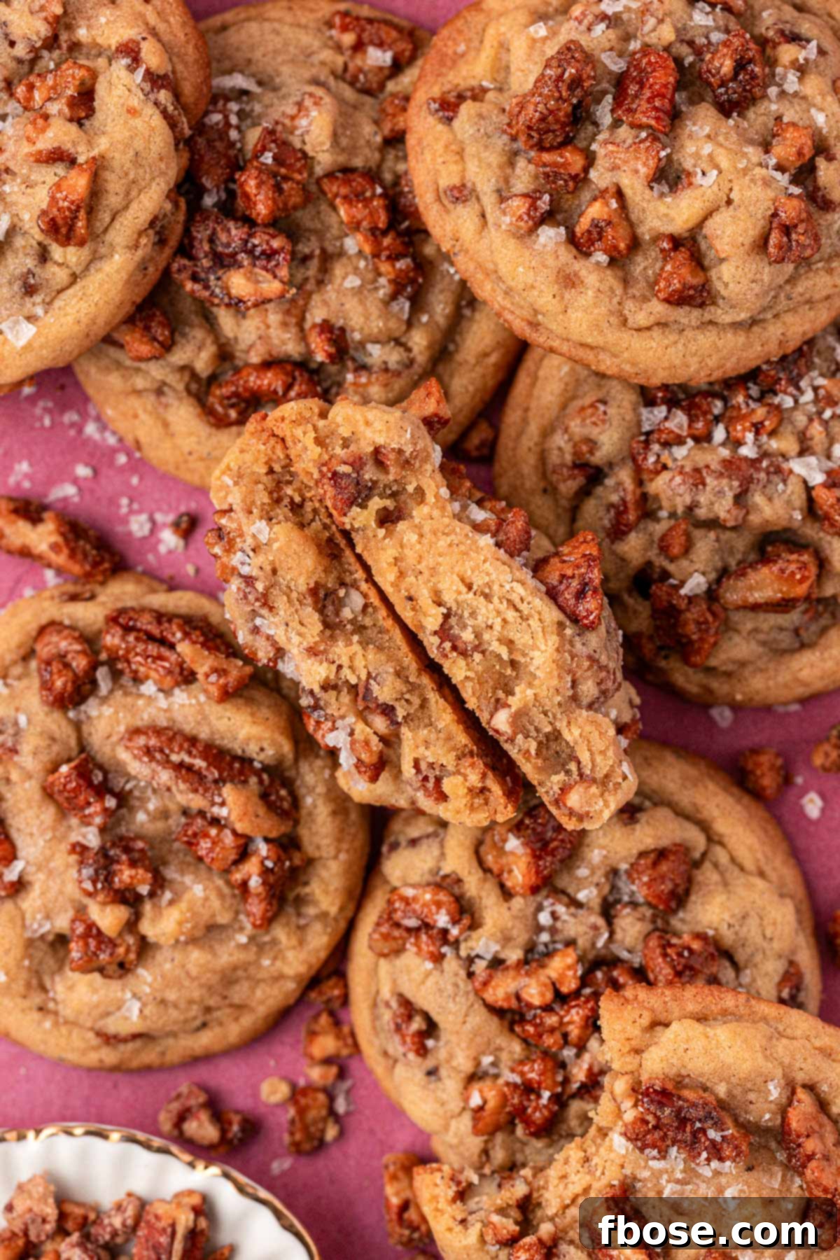 An overhead shot of freshly baked maple pecan cookies, some on a wire rack and some on a maroon surface, glistening with vanilla simple syrup and sea salt.