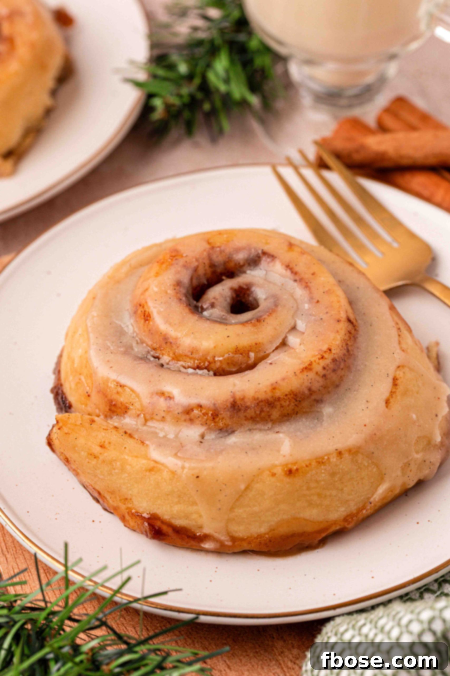 Eggnog Delight Swirls 15 A single eggnog cinnamon roll on a white plate with a gold fork, highlighting its perfect glaze and tempting swirls.